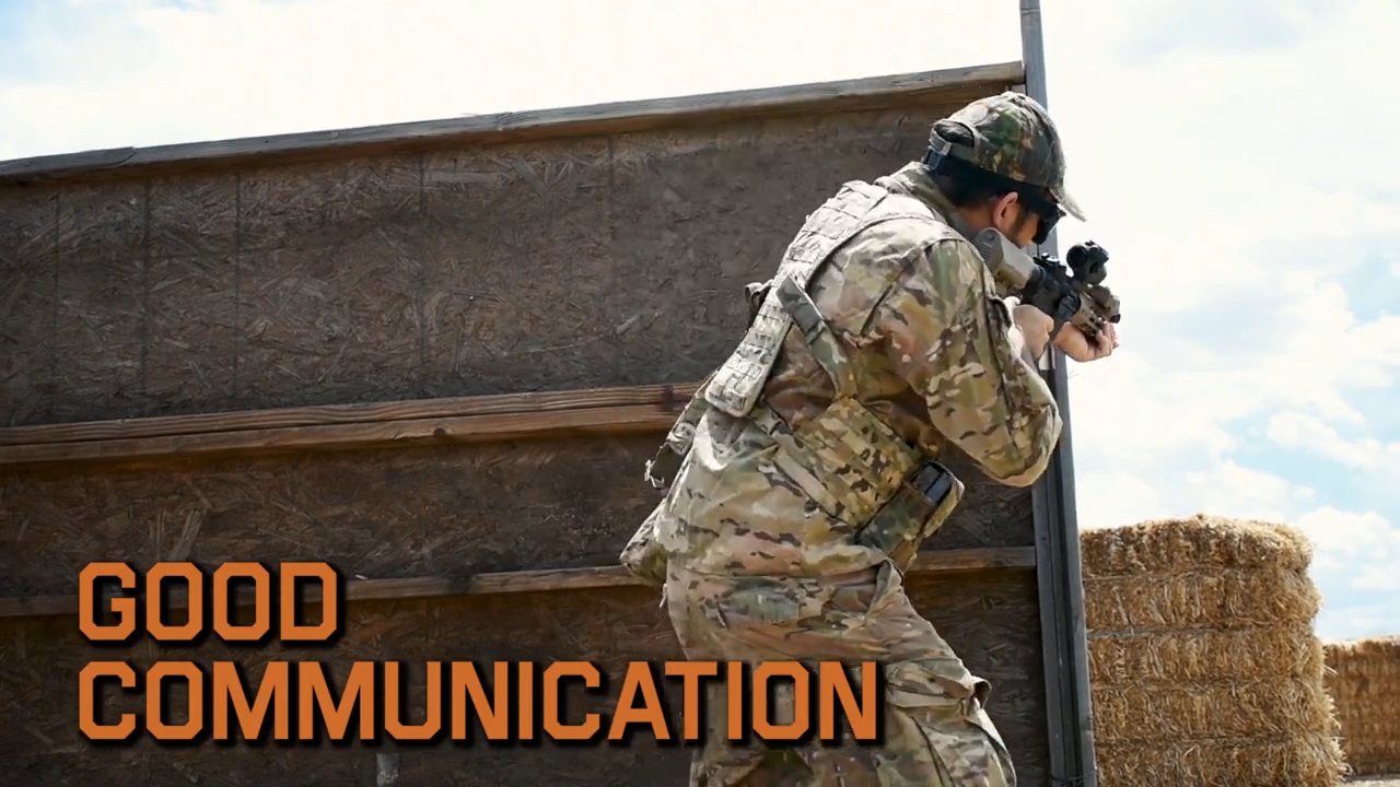 Airsoft player in multicam attire aiming a rifle beside a wooden barrier with the text "GOOD COMMUNICATION" showcasing effective teamwork in a tactical scenario.