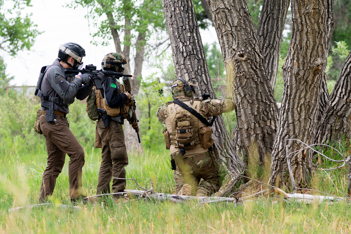 Three airsoft players in tactical gear engaging in a skirmish in a wooded area, using trees as cover.