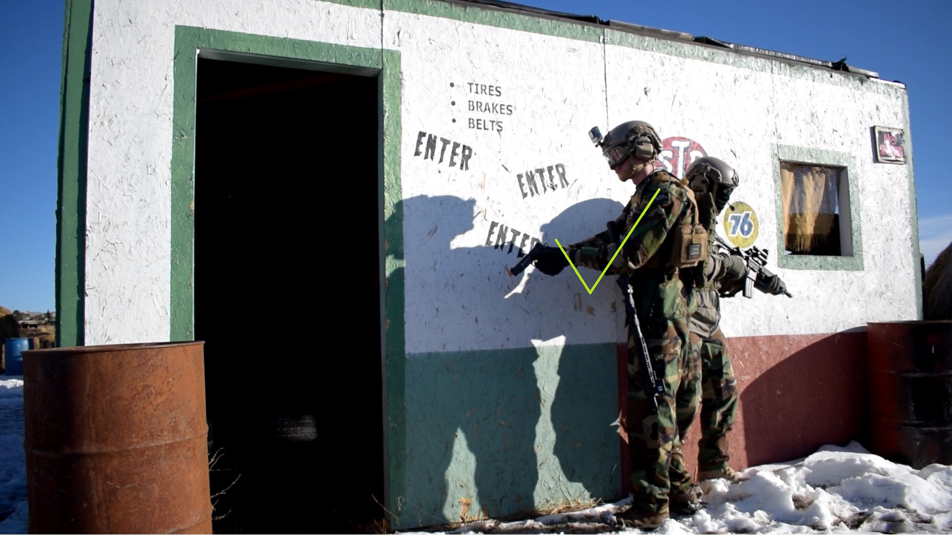 Two airsoft players in camouflage standing by a building with one player signaling to another, with a green check mark indicating correct action or communication.