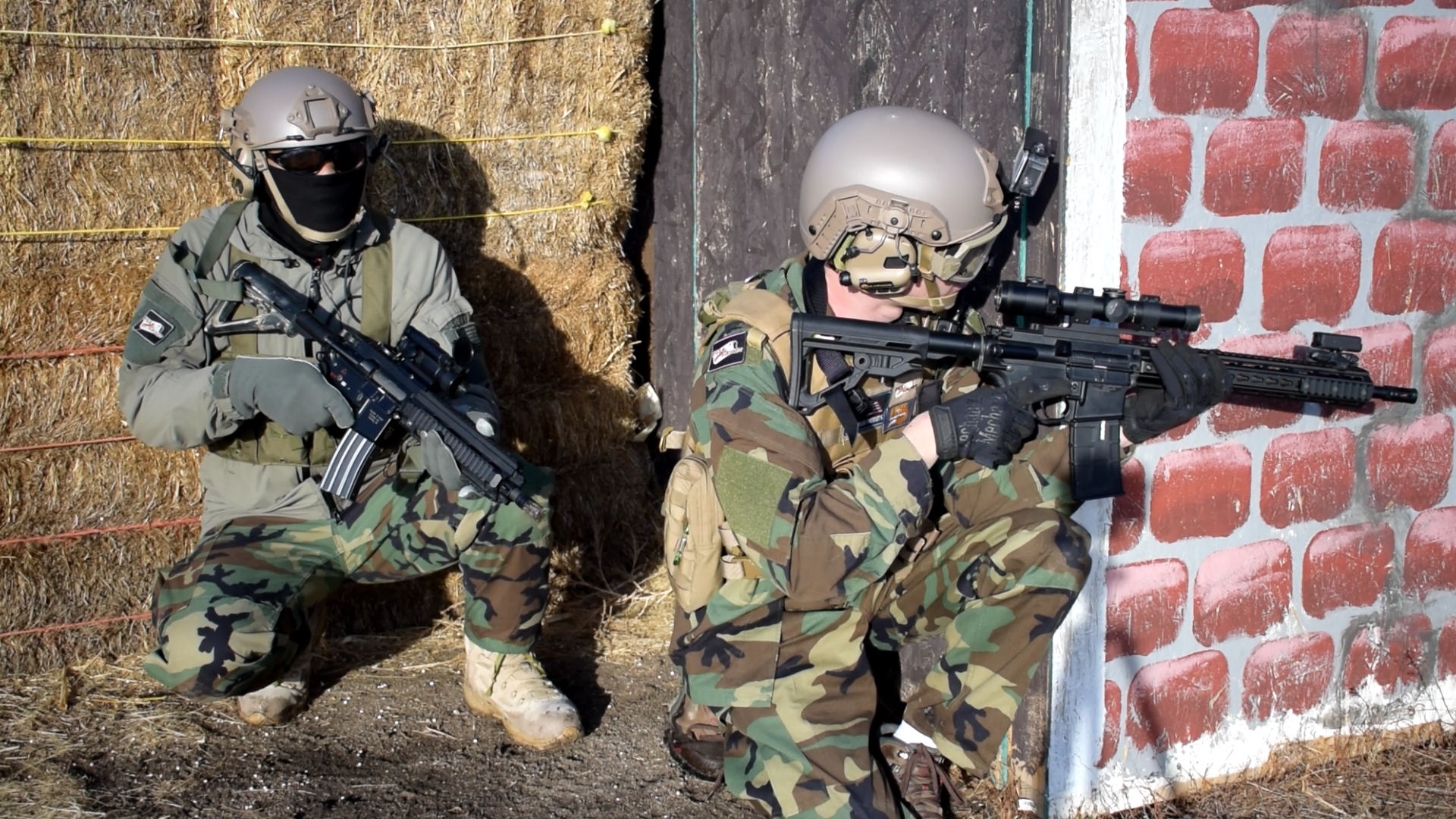 Two airsoft players in tactical gear and helmets taking cover by a corner of a brick wall and hay bales, ready with their rifles for engagement.