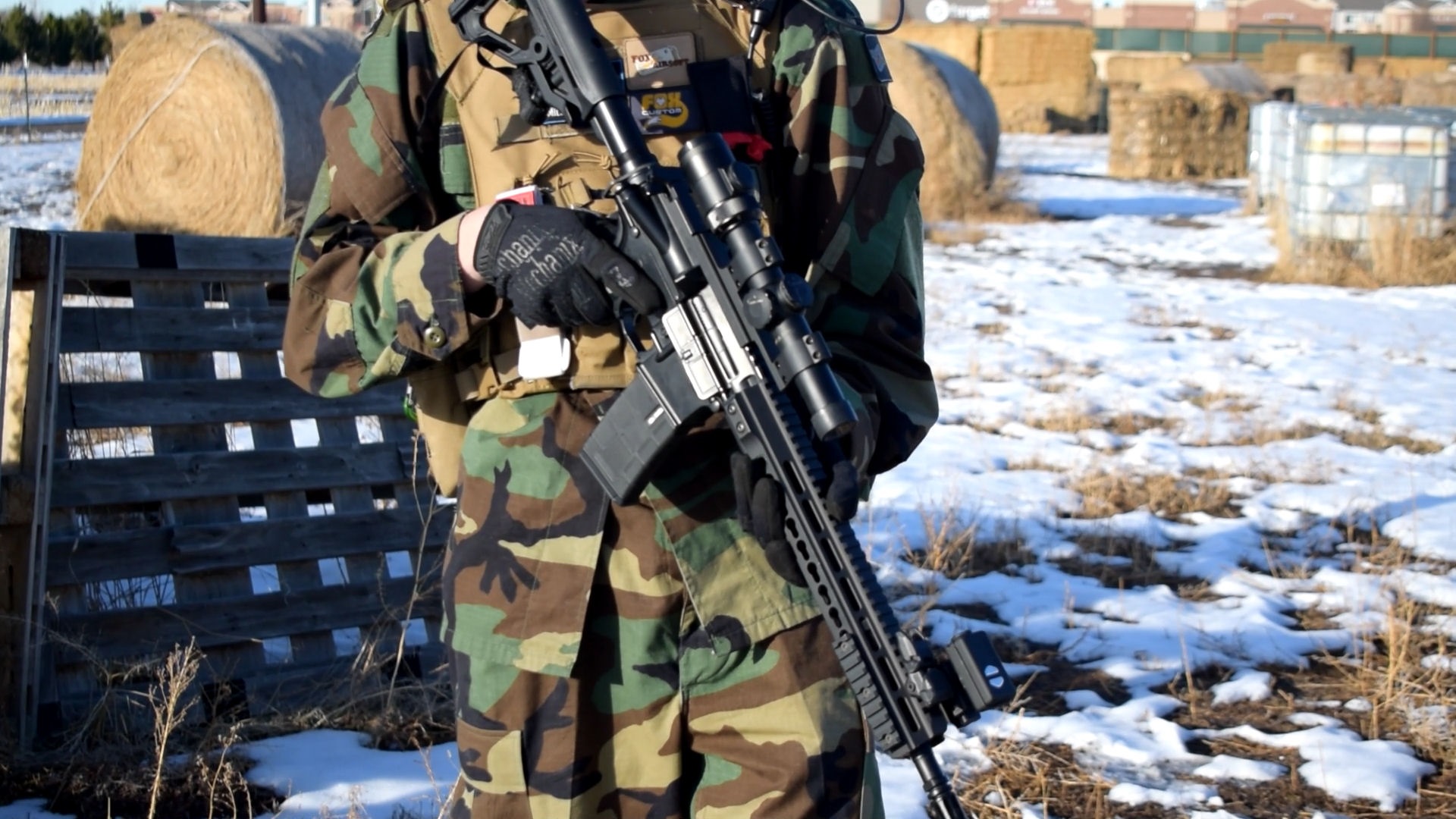 An airsoft player in camouflage uniform holding a rifle with optical sight, standing in a field with hay bales and patches of snow in the background.