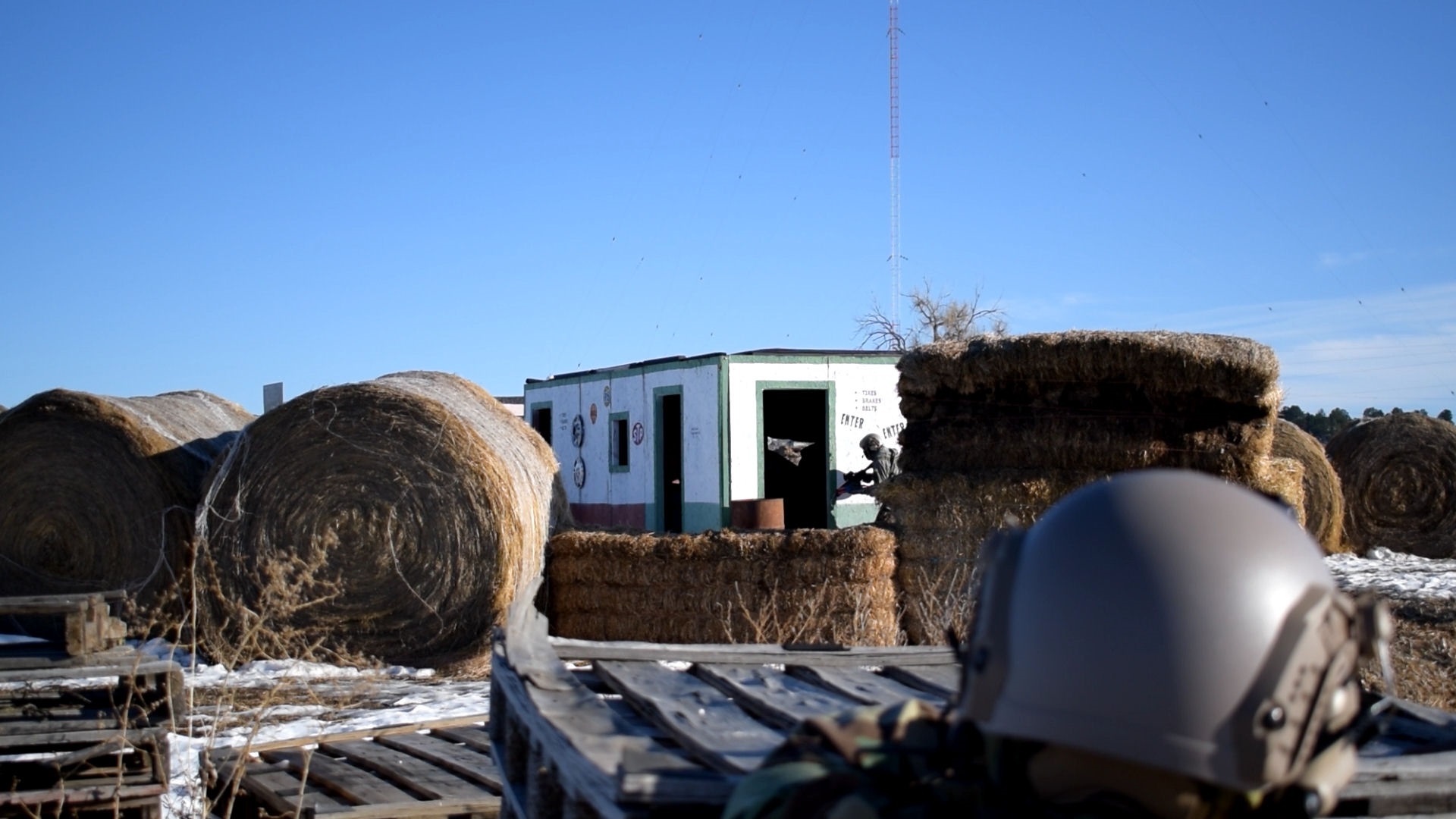 Airsoft player in foreground with helmet looking towards a mock building with target symbols, hay bales and clear blue sky in the background.