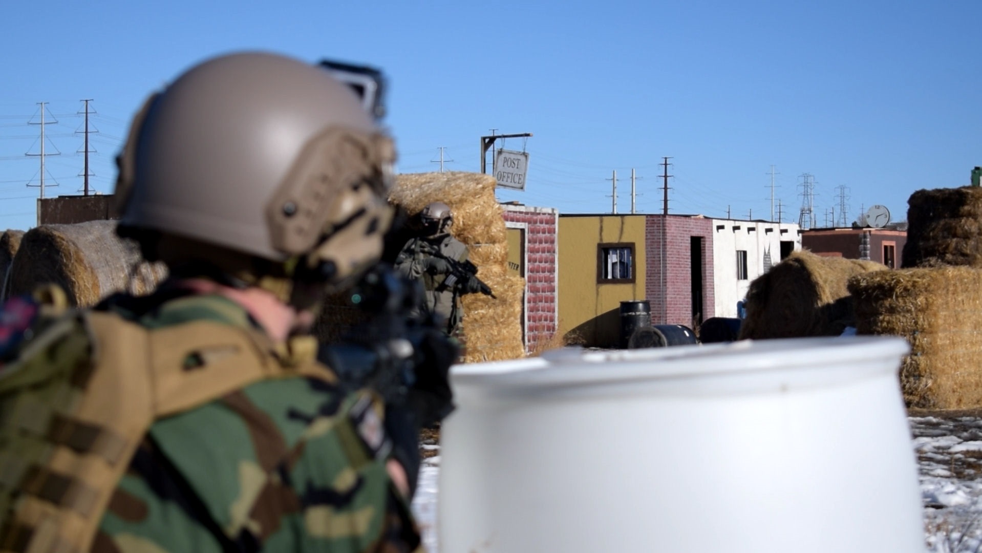 Airsoft player in combat gear observing through rifle scope with blurred foreground, urban landscape and hay bales in the background.