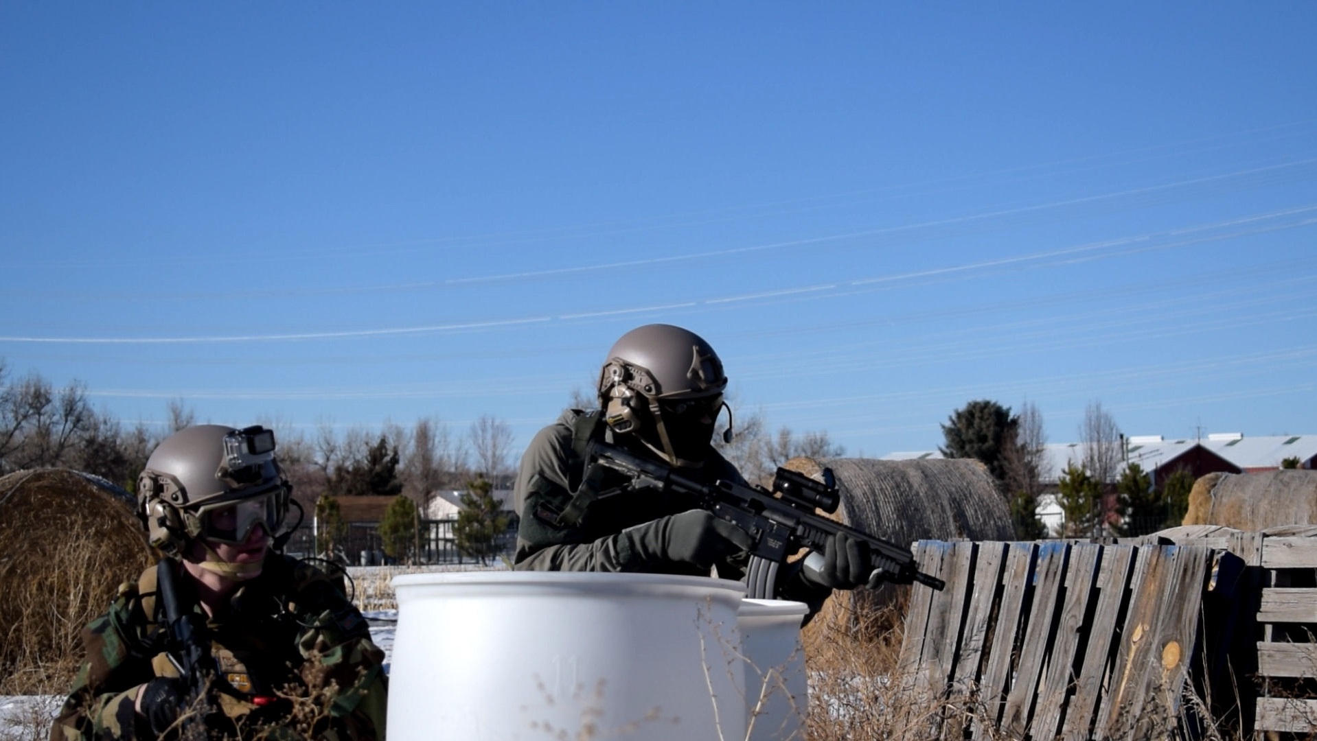 Two airsoft players in helmets and tactical gear taking positions behind cover in an outdoor field with clear skies above.