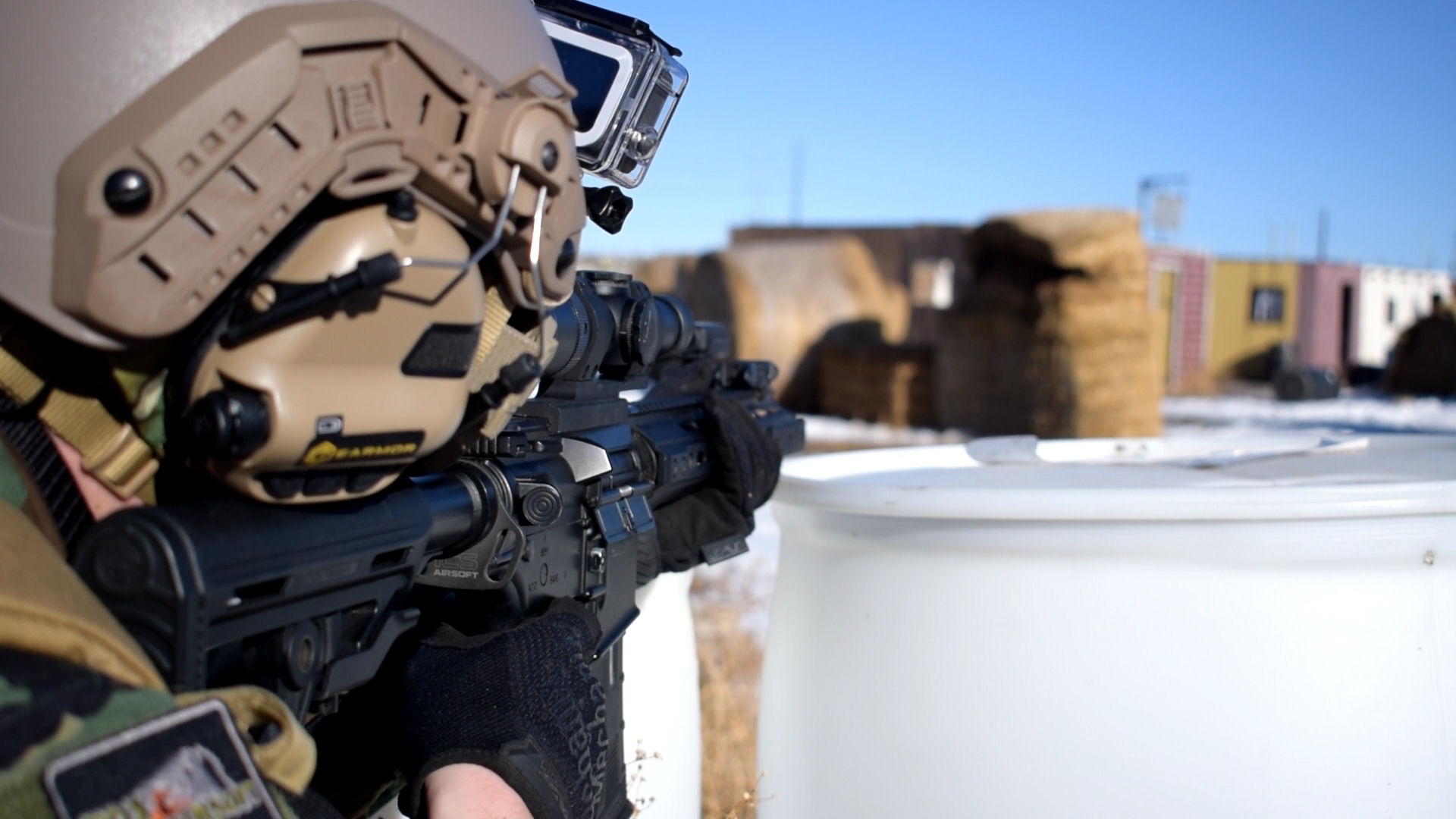 Airsoft player aiming down the sights of a rifle with an attached scope, wearing a helmet with communication gear and a mounted camera, against a backdrop of hay bales and urban buildings.