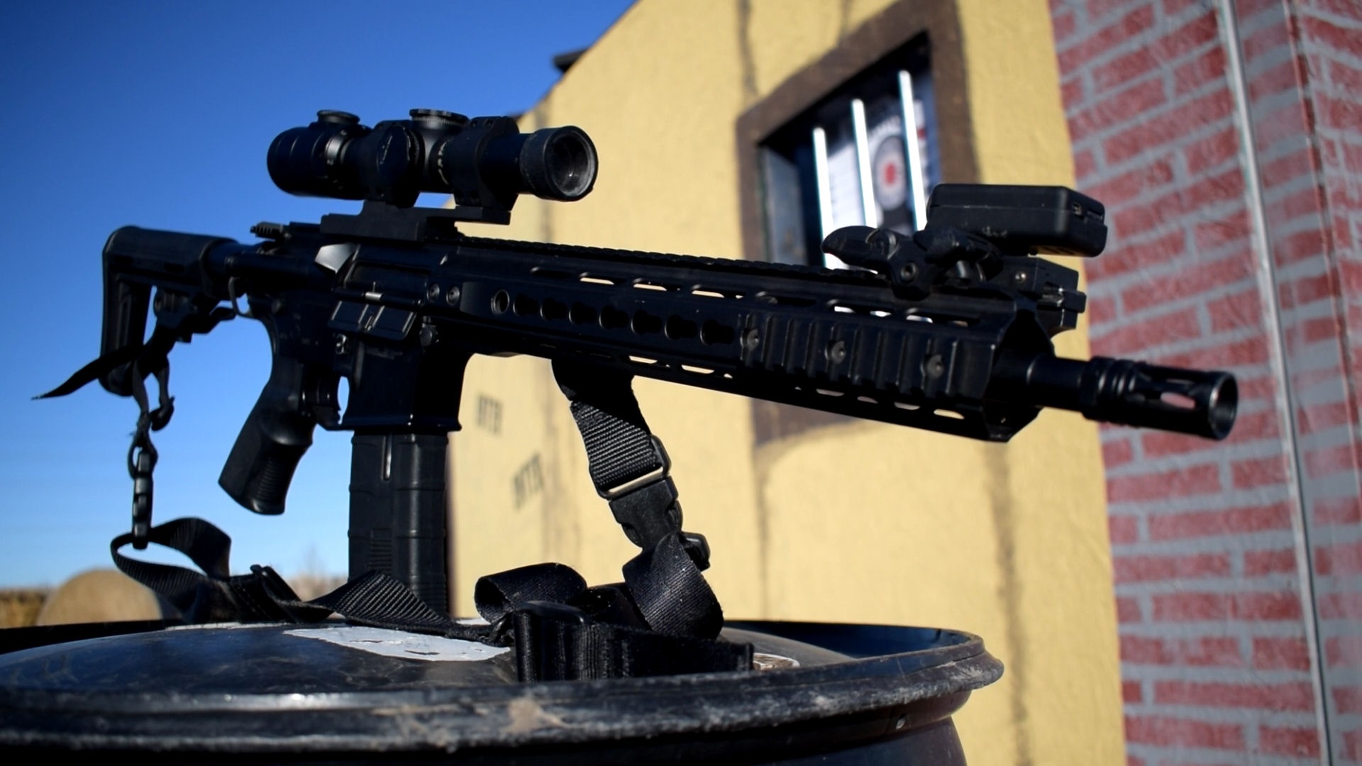 An airsoft rifle with a scope mounted on a stand, against the backdrop of a building with a brick wall design.