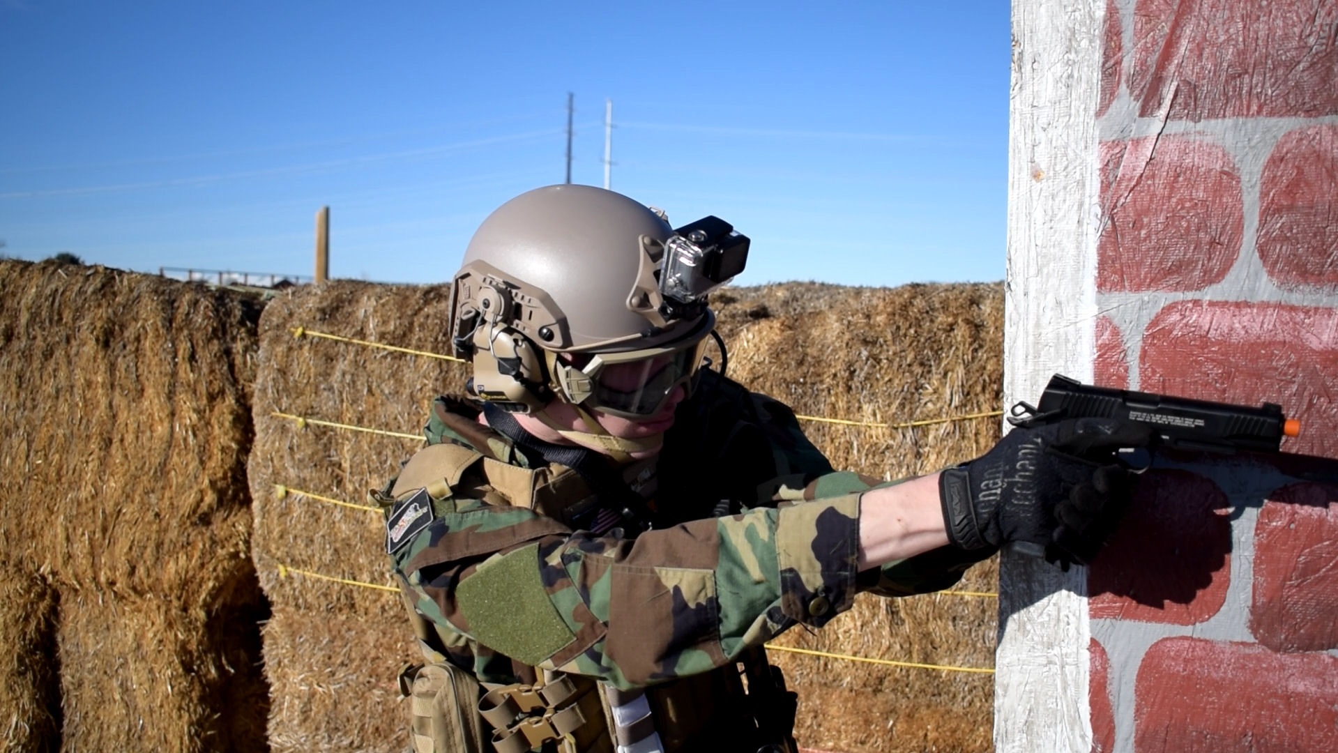 An airsoft player in a camouflage uniform and tactical helmet with a mounted camera, aiming a handgun beside a brick wall with hay bales in the background.