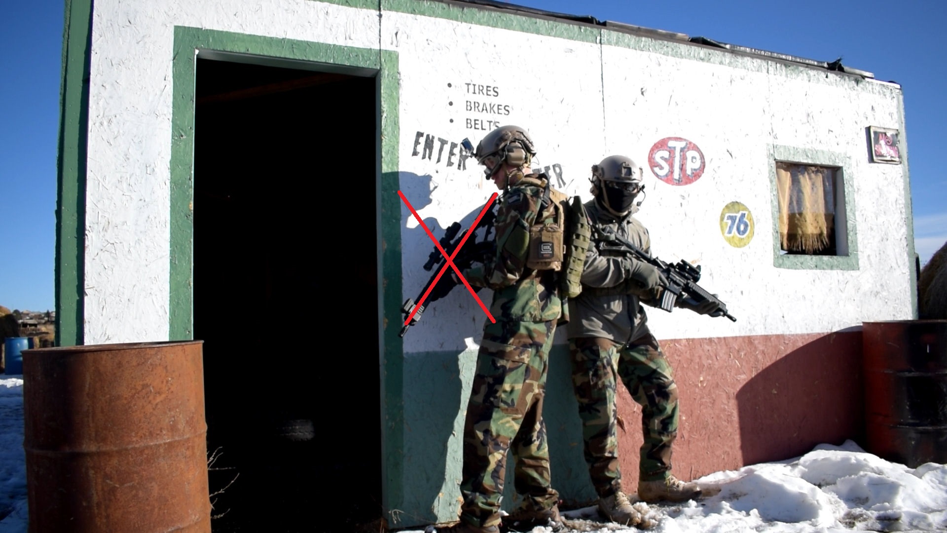 Two airsoft players in full combat gear standing against a wall with a red "X" over one player to possibly denote an instructional scenario or a safety violation.