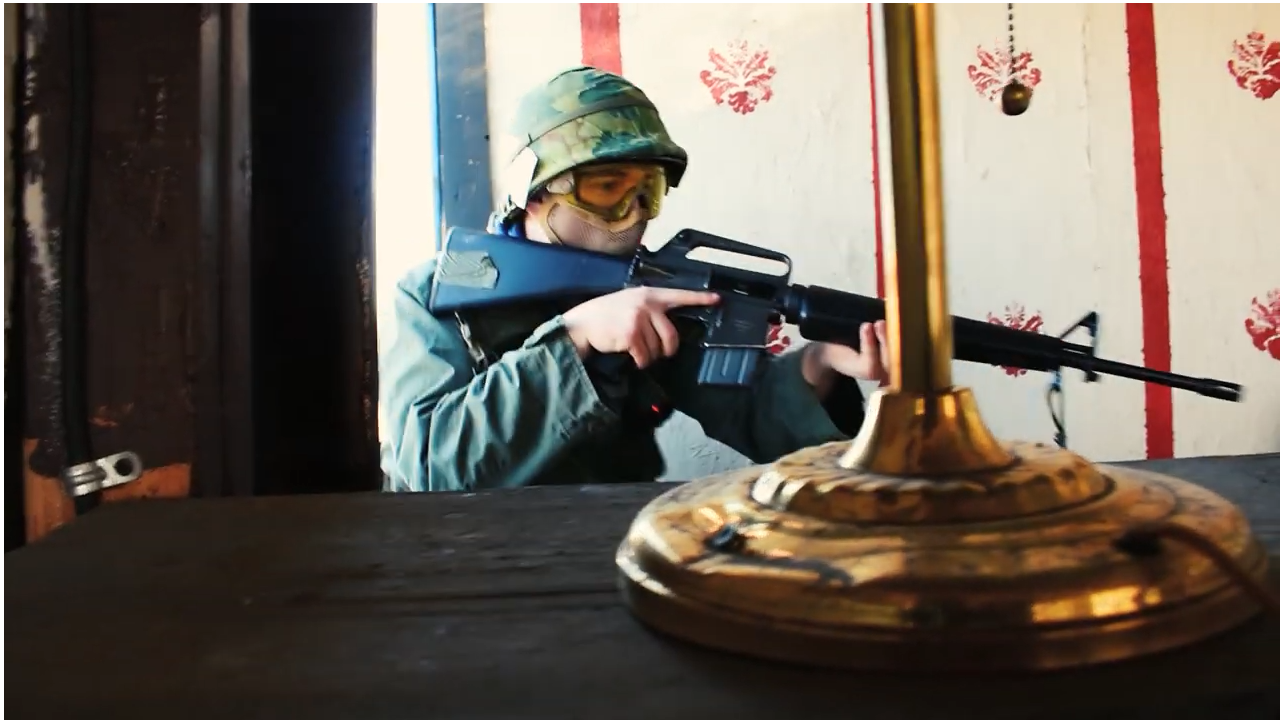 Airsoft player in full gear aiming a compact airsoft gun, with eye protection, in a staged indoor environment with striped wallpaper.