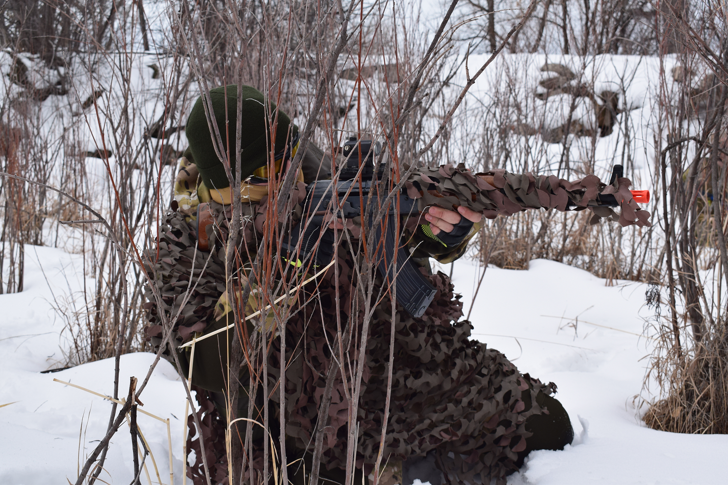 Airsoft sniper in a ghillie suit crouching in a snowy environment, blending into the surroundings while aiming a rifle.