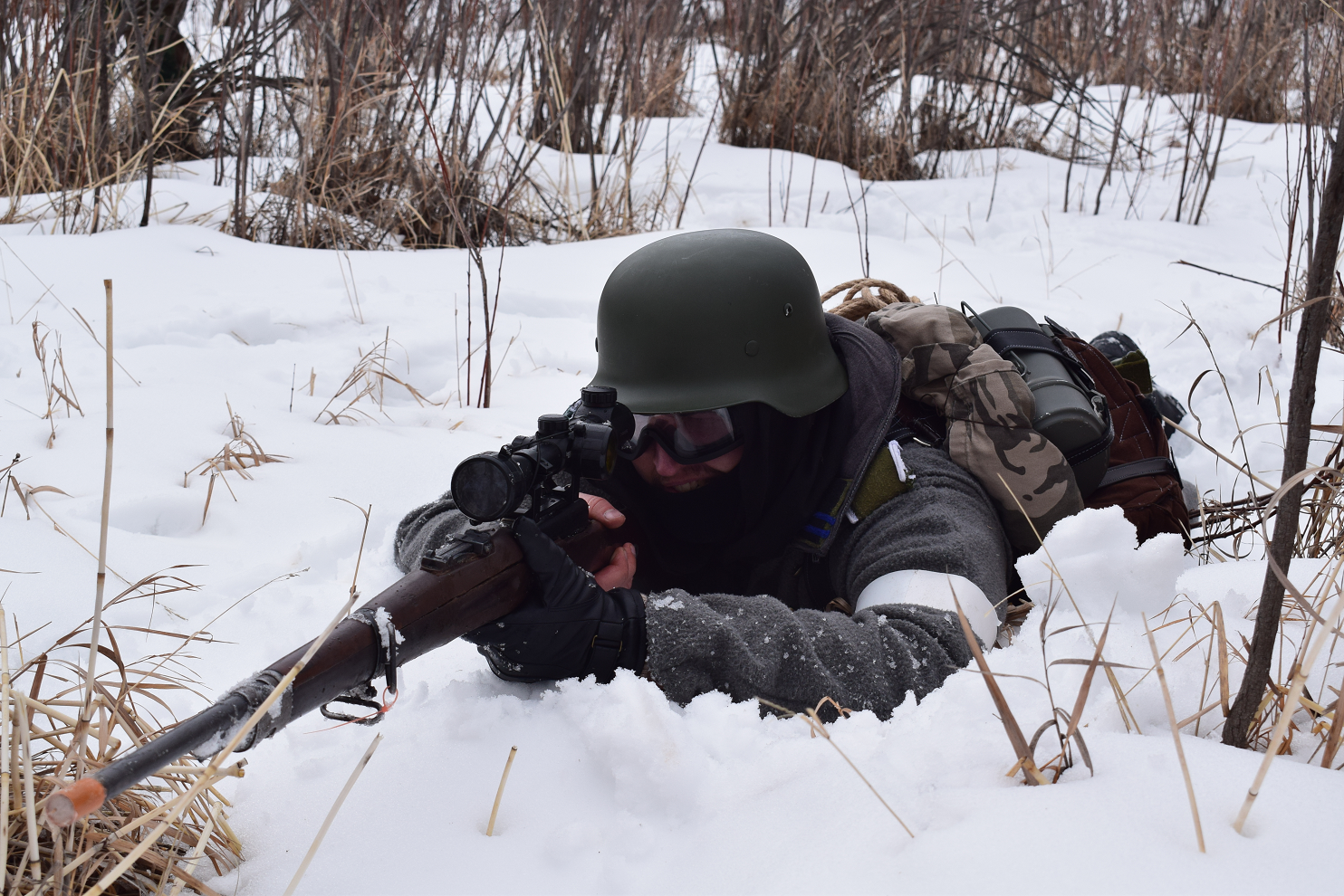 Airsoft player lying prone in the snow, aiming with a scoped rifle, wearing winter camouflage and protective gear.