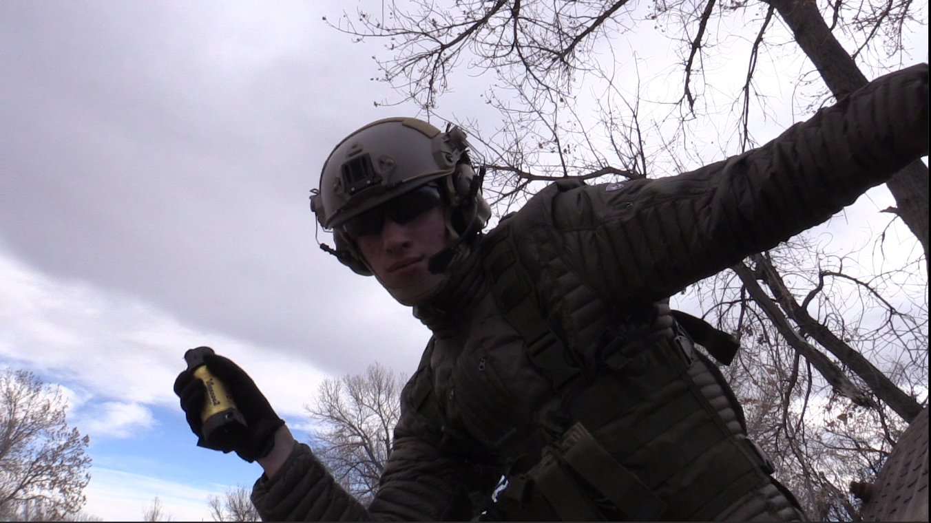 Airsoft player in tactical gear holding a yellow smoke grenade, preparing to deploy it during a game in a wooded area.